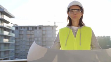 Kadın bir mimar ya da mühendis, güvenlik kasketi ve yelek giyiyor. Şafak vakti inşaat alanının planını tutuyor. Mimarlık ve mühendislik kavramları.