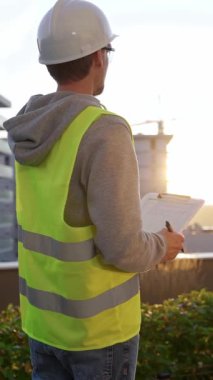 Beyaz kasklı ve güvenlik yeleği olan mühendis, sabahın erken saatlerinde dikey olarak bir inşaat alanını incelerken bir panoya not alıyor. Mimarlık ve mühendislik