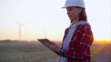 Kadın mühendis, güneş batarken rüzgâr türbinleriyle dolu bir alanda bir panoya not alıyor. Temiz enerji ve mühendislik denetimi kavramı.