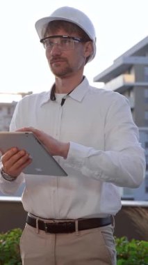 Beyaz gömlekli ve kasklı bir inşaat mühendisi güneş doğarken, ön ve dikey manzaralı bir inşaat alanını incelerken dijital tablet kullanıyor. Mimarlık konsepti.