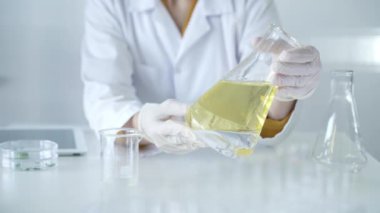 Beyaz koruyucu eldivenler ve laboratuvar önlüğü giyen tanınmayan bilim adamı laboratuvardaki masada otururken elinde sarı yağ sıvısı bulunan bir deney şişesi tutuyor. Tıp ve Bilim.