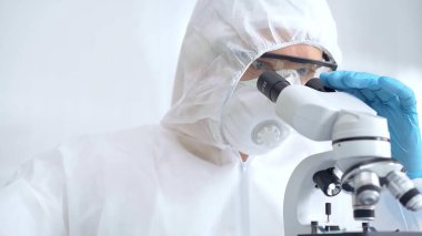 Laboratuvar ortamında mikroskop kullanarak örnekleri analiz eden bilim adamı. Laboratuvar önlüğü ve koruyucu ekipman takmış odaklanmış bir araştırmacının yakın çekimi..