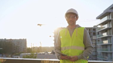 İnşaat şantiyesi mühendisi elinde tablet tutuyor ve arka planda gündoğumuyla birlikte yelek ve şapka giyiyor. Portre videosu.