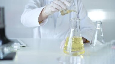 Laboratuvar önlüğü ve beyaz koruyucu eldiven giyen bir bilim adamı, laboratuarda bir şişeden diğerine sarı yağlı bir sıvı döküyor, kapatın. Tıp ve bilim konsepti.
