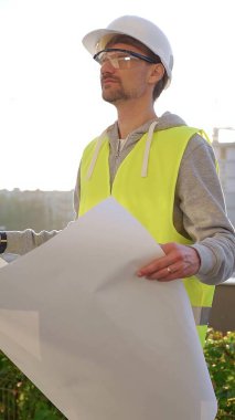Erkek ve kadın mimarlar veya mühendisler güvenlik kasketleri ve yelekleri giyip şemaları tutuyorlar ve gün doğumunda inşaat alanında bir şey tartışıyorlar, ön dikey görüş. Mimarlık konsepti.