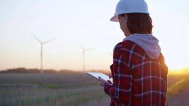 Kadın mühendis, güneş batarken rüzgar türbinleriyle dolu bir alanda tablet bilgisayarla not alıyor. Temiz enerji ve mühendislik kavramı.