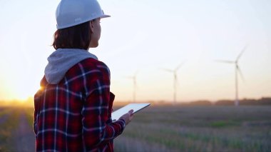 Kadın mühendis, güneş batarken rüzgar türbinleriyle dolu bir alanda tablet bilgisayarla not alıyor. Temiz enerji ve mühendislik kavramı.