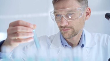 Koruyucu gözlüklü bilim adamı mavi bir sıvıyla laboratuvar tüpünü inceliyor. Mikrobiyoloji ve bilim konsepti.
