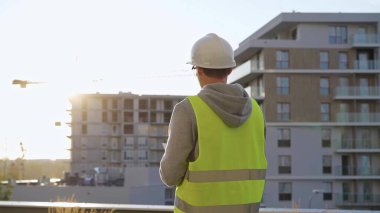 Beyaz şapka ve güvenlik yeleği giyen inşaat mühendisi, sabahın erken saatlerinde bir inşaat alanını incelerken tablet bilgisayar kullanıyor..