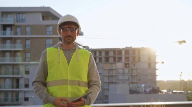İnşaat şantiyesi mühendisi elinde tablet tutuyor ve arka planda gündoğumuyla birlikte yelek ve şapka giyiyor. Portre videosu.