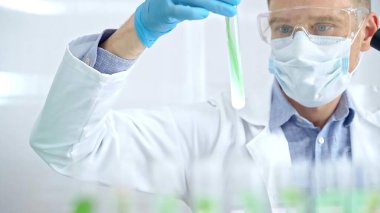 Koruyucu eldivenleri ve gözlükleri olan bir bilim adamı yeşil sıvıyla laboratuvar tüpünü inceliyor. Mikrobiyoloji ve bilim konsepti.