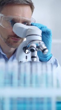 Koruyucu gözlüklü ve eldivenli bilim adamı laboratuarda mikroskop kullanıyor, araştırmacının dikey portresi. Mikrobiyoloji bilim konsepti.