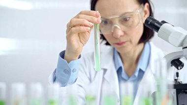 Koruyucu gözlüklü kadın bilim adamı yeşil sıvıyla laboratuvar tüpünü inceliyor. Mikrobiyoloji ve bilim konsepti.