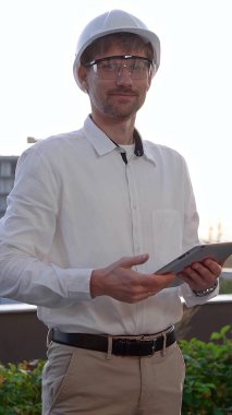Beyaz gömlekli ve başlıklı bir mimari mühendisi güneş doğarken, ön ve dikey görünümde bir inşaat alanını incelerken dijital tablet kullanır..