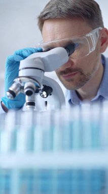 Koruyucu gözlüklü ve eldivenli bilim adamı laboratuarda mikroskop kullanıyor, araştırmacının dikey portresi. Mikrobiyoloji bilim konsepti.