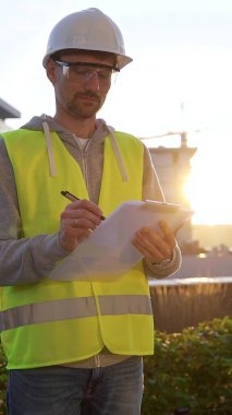 Beyaz kasketli ve güvenlik yeleği olan mühendis güneş doğarken bir inşaat alanını dikey olarak incelerken not alıyor. Mimarlık ve mühendislik.