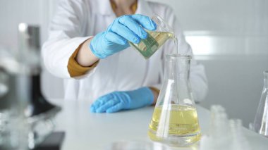 Laboratuvar önlüğü ve mavi koruyucu eldiven giyen bir bilim adamı laboratuarda bir şişeden diğerine sarı bir sıvı boşaltıyor. Tıp ve bilim kavramları.