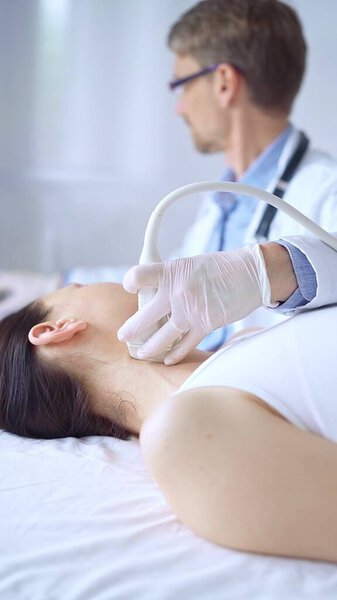 Врач мужчина в белых защитных перчатках использует ультразвуковое оборудование на шее пациентки, лежащей на обследовании. Концепция медицины.