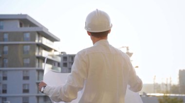 Güvenlik kaskı ve gözlük takan inşaat alanı yöneticisi, güneş doğarken veya batarken elinde planlar tutuyor ve bir inşaat alanını inceliyor. Mimarlık ve mühendislik.