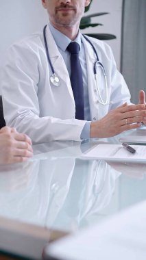 Doctor consulting patient. Male physician is gesturing above a clipboard while talking with a woman in a clinic, close up. Medicine concept. clipart