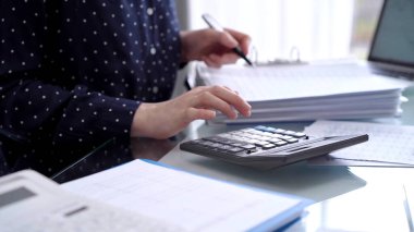 Businesswoman using calculator and reviewing ring folder of financial documents with magnifying glass at desk in modern office. Audit and taxes in business. clipart