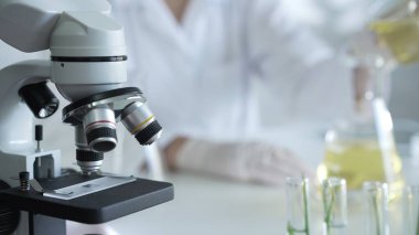 Laboratuvar önlüğü ve beyaz koruyucu eldiven giyen bir bilim adamı, laboratuara yakın mikroskoba sarı yağlı bir sıvıyı döküyor. Tıp ve bilim konsepti.