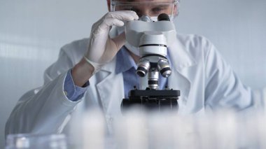 Beyaz eldiven, koruyucu gözlük ve tıbbi maske takan bilim adamı araştırmacı modern laboratuarda mikroskop kullanıyor, portre görüşü. Gösterişsiz ışıklandırma fotoğrafı. Bilim ve Tıp.