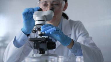 Laboratuvar önlüğü, beyaz koruyucu eldiven ve gözlük giyen kadın bilim adamı modern laboratuvar düzeninde mikroskop kullanıyor, portre görüntüsü. Gösterişsiz ışıklandırma fotoğrafı. Bilim ve Tıp.
