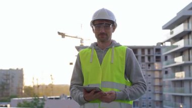 İnşaat şantiyesi mühendisi elinde tablet tutuyor ve sabah erken saatlerde arka planda güneş doğarken sıkı şapka ve güvenlik yeleği giyiyor..