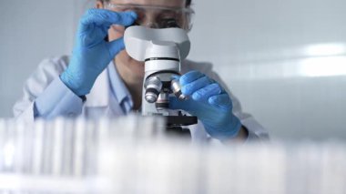 Laboratuvar önlüğü, beyaz koruyucu eldiven ve gözlük giyen kadın bilim adamı modern laboratuvar düzeninde mikroskop kullanıyor, portre görüntüsü. Çaktırmadan ışıklandırma videosu. Bilim ve Tıp.