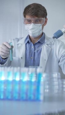 Laboratuvar önlüğü, beyaz tıbbi eldiven, maske ve koruyucu gözlük giyen erkek bilim adamı araştırmacı modern laboratuvarda mikropipet doldurma tüpleri kullanıyor. Çaktırmadan ışıklandırma videosu. Bilim konsepti.