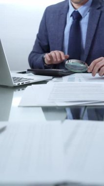 Mavi takım elbiseli bir işadamı büyüteçle mali raporları analiz ediyor ve profesyonel ofis ortamında hesap makinesi kullanıyor. Düşük ışıklandırma, yakın dikey görüntü. İş adamları, denetim.