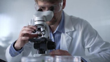 Laboratuvar önlüğü giyen ve koruyucu gözlük takan erkek bilim adamı mikroskop kullanarak modern laboratuvar düzeninde araştırma yapıyor, portre görüntüsü çiziyor. Çaktırmadan ışıklandırma videosu. Bilim ve Tıp Konsepti.