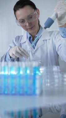 Laboratuvar önlüğü, beyaz tıbbi eldiven ve koruyucu gözlük giyen kadın bilim adamı modern laboratuarda mikropipet doldurma tüpleri kullanıyor. Çaktırmadan ışıklandırma videosu. Bilim konsepti.
