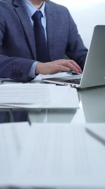 Mavi takım elbiseli iş adamı ofis setindeki cam masada dizüstü bilgisayar, hesap makinesi ve belgelerle çalışıyor. Düşük ışıklandırma, yakın dikey görüntü. İş adamları ve denetim.