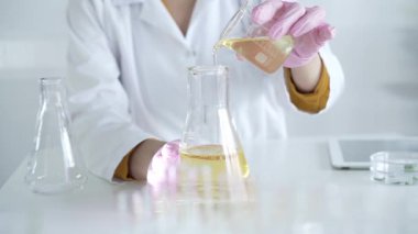 Laboratuvar önlüğü ve pembe koruyucu eldiven giyen bir bilim adamı laboratuarda sarı yağlı bir sıvıyı laboratuvardan diğerine döküyor. Tıp ve Bilim.