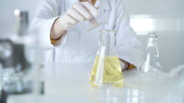 Laboratuvar önlüğü ve beyaz koruyucu eldiven giyen bir bilim adamı, laboratuarda bir şişeden diğerine sarı yağlı bir sıvı döküyor, kapatın. Tıp ve bilim konsepti.