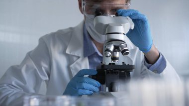 Mavi eldiven, koruyucu gözlük ve tıbbi maske takan erkek bilim adamı araştırmacı modern laboratuarda mikroskop kullanıyor, portre görüşü. Gösterişsiz ışıklandırma fotoğrafı. Bilim ve Tıp.