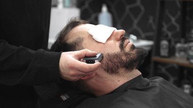 Profesyonel berber, sakin bir müşterinin sakalını tıraş ederken ustura kullanır, modern bir berber dükkanında kaliteli bir tımar deneyimi sağlar.