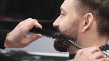 Profesyonel berber, müşterinin sakalını makas ve tarak ile tarayıp keserek modern bir berber dükkanında şık ve titiz bir tımar deneyimi sağlar.