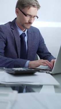 Mavi takım elbiseli iş adamı ofis setindeki cam masada dizüstü bilgisayar, hesap makinesi ve belgelerle çalışıyor. Düşük ışıklandırma, dikey portre. İş adamları ve denetim.