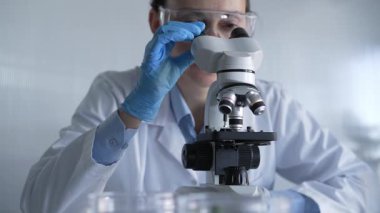 Laboratuvar önlüğü, beyaz koruyucu eldiven ve gözlük giyen kadın bilim adamı modern laboratuvar düzeninde mikroskop kullanıyor, portre görüntüsü. Çaktırmadan ışıklandırma videosu. Bilim ve Tıp.