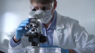 Mavi eldiven, koruyucu gözlük ve tıbbi maske takan bilim adamı araştırmacı modern laboratuarda mikroskop kullanıyor, portre görüntüsü. Çaktırmadan ışıklandırma videosu. Bilim ve Tıp.