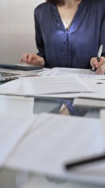Mavi elbise giyen iş kadını profesyonel ofis ortamında dizüstü bilgisayar, hesap makinesi ve belgelerle çalışıyor. Düşük ışıklandırma, yakın dikey görüntü. İş adamları, denetim kavramı.