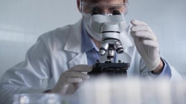 Beyaz eldiven, koruyucu gözlük ve tıbbi maske takan bilim adamı araştırmacı modern laboratuarda mikroskop kullanıyor, portre görüşü. Çaktırmadan ışıklandırma videosu. Bilim ve Tıp.