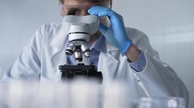 Mavi eldiven, koruyucu gözlük ve tıbbi maske takan bilim adamı araştırmacı modern laboratuarda mikroskop kullanıyor, portre görüntüsü. Çaktırmadan ışıklandırma videosu. Bilim ve Tıp.