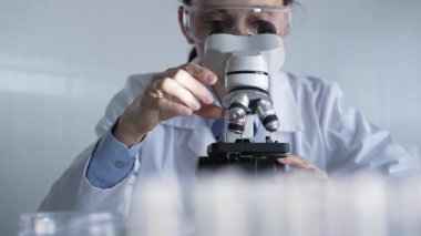 Laboratuvar önlüğü ve koruyucu gözlük takan kadın bilim adamı modern laboratuvar düzeneğinde mikroskop kullanıyor. Düşük tuşlu ışıklandırma videosu.