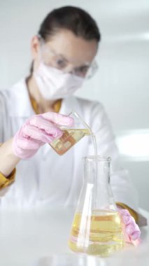 Laboratuvar önlüğü, pembe eldiven ve koruyucu gözlük takan kadın bilim adamı deney şişesinden Erlenmeyer şişesine laboratuvar, portre ve dikey görüntüye sarı bir sıvı döküyor. Tıp, bilim..