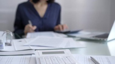 Beyaz hesap makinesi, kalem ve belgeler muhasebecilerin masasında yatıyor. Mavi elbise giyen iş kadını arka planda belgelerle çalışıyor. İş ve denetim kavramı.