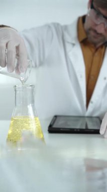 Laboratuvar önlüğü, beyaz eldiven ve koruyucu gözlük takan bilim adamı laboratuvar ortamında sarı yağlı sıvıyı bir bardaktan diğerine döküyor. Bilim ve tıp kavramları.